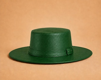 May include: A dark green flat-brimmed hat with a structured cylindrical crown and a matching green band featuring a small rectangular buckle detail. The hat has a smooth, uniform finish and a wide, circular brim that extends from the base.