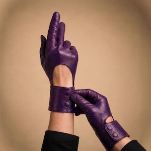May include: A pair of deep purple leather gloves with a wrist strap and button closure. The gloves have a cutout detail on the back of the hand. The gloves are shown against a neutral background.