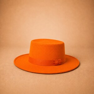 May include: A vibrant orange flat-brimmed hat featuring a structured cylindrical crown and a wide, circular brim. The hat is accented with a matching orange ribbon band that encircles the base of the crown, creating a uniform and solid colour.