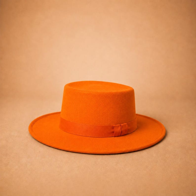 May include: A vibrant orange flat-brimmed hat featuring a structured cylindrical crown and a wide, circular brim. The hat is accented with a matching orange ribbon band that encircles the base of the crown, creating a uniform and solid colour.