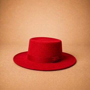 May include: A vibrant red hat featuring a flat crown and a wide, circular brim. The hat is constructed from a smooth, felt-like material and includes a matching red ribbon band wrapped around the base of the crown, adding a subtle texture.
