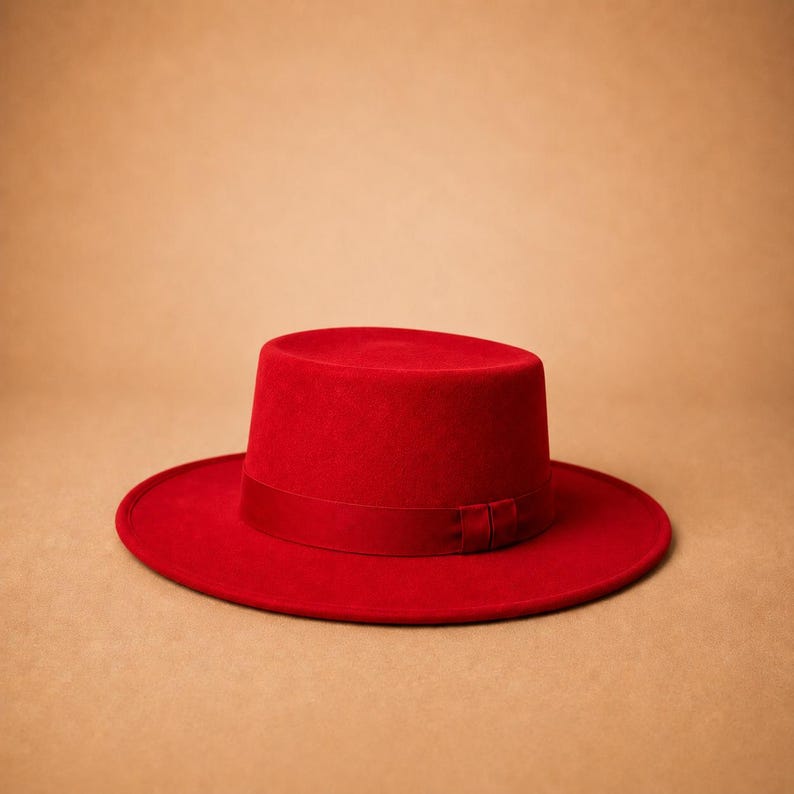May include: A vibrant red hat featuring a flat crown and a wide, circular brim. The hat is constructed from a smooth, felt-like material and includes a matching red ribbon band wrapped around the base of the crown, adding a subtle texture.