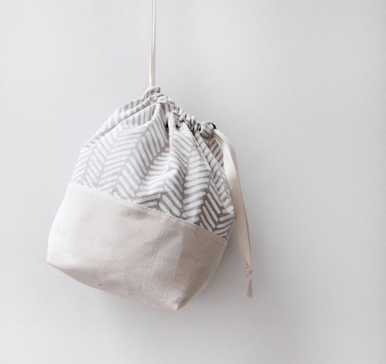 Puede incluir: Una bolsa de cord&oacute;n de color gris y blanco con un fondo beige. La bolsa est&aacute; colgada de una cuerda blanca.