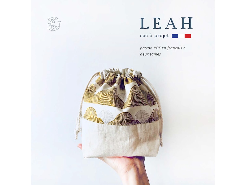 May include: A beige drawstring bag with a gold and white patterned fabric. The bag is labeled "LEAH sac &agrave; projet" and features a French flag.