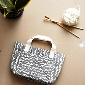 May include: A grey and white tote bag with a woven pattern. The bag has two handles and is sitting on a beige surface. There are knitting needles and a ball of yarn in the background.