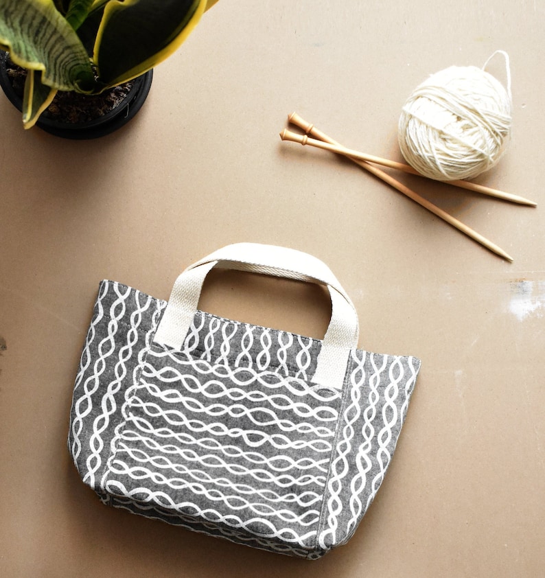May include: A grey and white tote bag with a woven pattern. The bag has two handles and is sitting on a beige surface. There are knitting needles and a ball of yarn in the background.