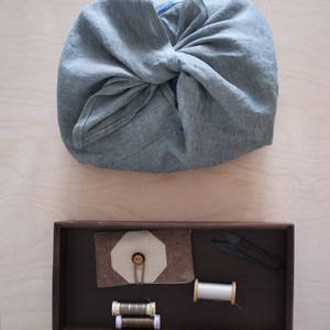 May include: A gray linen fabric bundle tied in a knot, above a dark wooden tray. The tray contains sewing supplies: thread spools, a fabric button holder, and small scissors. The background is a light wood surface.