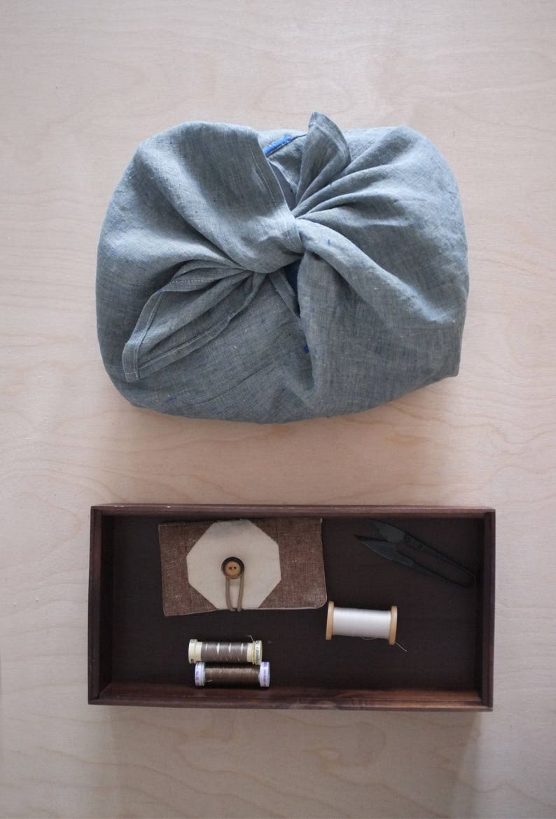 May include: A gray linen fabric bundle tied in a knot, above a dark wooden tray. The tray contains sewing supplies: thread spools, a fabric button holder, and small scissors. The background is a light wood surface.