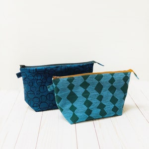 May include: Two fabric pouches with zippers. One is dark blue with a black circle pattern, and the other is teal with a green diamond pattern. Both have black zippers and are on a white wooden surface.