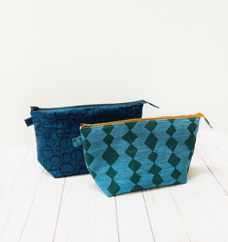 May include: Two fabric pouches with zippers. One is dark blue with a black circle pattern, and the other is teal with a green diamond pattern. Both have black zippers and are on a white wooden surface.