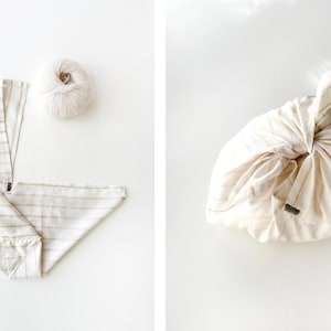 May include: Two-panel image showing a beige and white striped fabric, one unfolded and one tied into a bundle. A ball of beige yarn sits next to the unfolded fabric. The fabric is a reusable wrapping cloth.