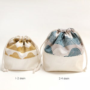 Puede incluir: Dos bolsas de cord&oacute;n hechas de tela blanca con una parte superior estampada. La bolsa m&aacute;s peque&ntilde;a tiene un estampado dorado y blanco y la bolsa m&aacute;s grande tiene un estampado azul y blanco. Las bolsas est&aacute;n etiquetadas como "1-2 ovillos" y "2-4 ovillos".