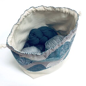 Puede incluir: Una bolsa de cord&oacute;n blanco con un patr&oacute;n geom&eacute;trico azul y blanco. La bolsa est&aacute; llena de ovillos de lana en tonos grises y azules.