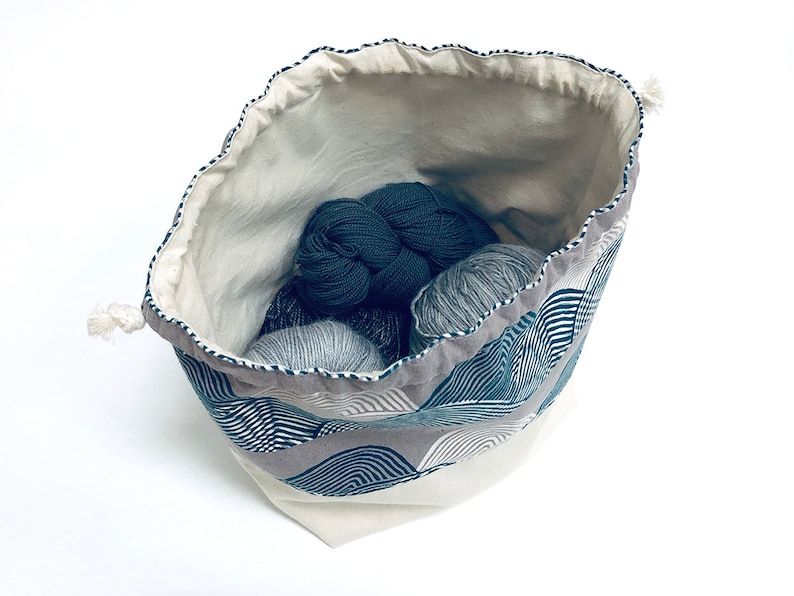 Puede incluir: Una bolsa de cord&oacute;n blanco con un patr&oacute;n geom&eacute;trico azul y blanco. La bolsa est&aacute; llena de ovillos de lana en tonos grises y azules.