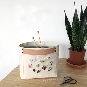 May include: A white canvas storage bin with a brown leather strap and a pocket on the front. The pocket is decorated with various pins and badges, including a cat, an avocado, a lobster, and a spool of thread. The bin is sitting on a wooden table next to a pair of scissors and a potted plant.