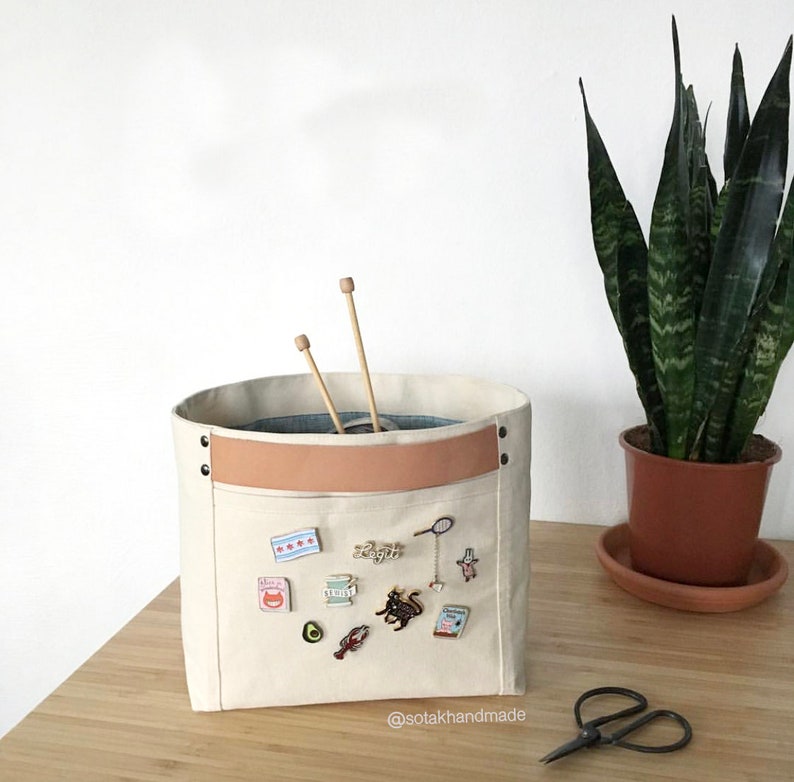 May include: A white canvas storage bin with a brown leather strap and a pocket on the front. The pocket is decorated with various pins and badges, including a cat, an avocado, a lobster, and a spool of thread. The bin is sitting on a wooden table next to a pair of scissors and a potted plant.