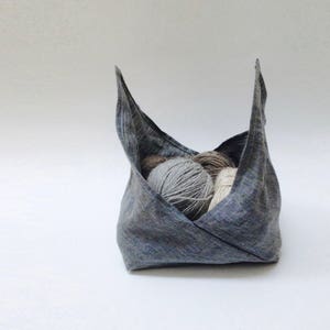 Puede incluir: Una bolsa de tela de lino gris con la parte superior doblada, que contiene tres ovillos de lana en tonos grises, marrones y blancos.