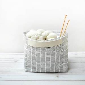 May include: A white fabric storage bin with a black and white geometric pattern. The bin is filled with white yarn balls and two wooden knitting needles.