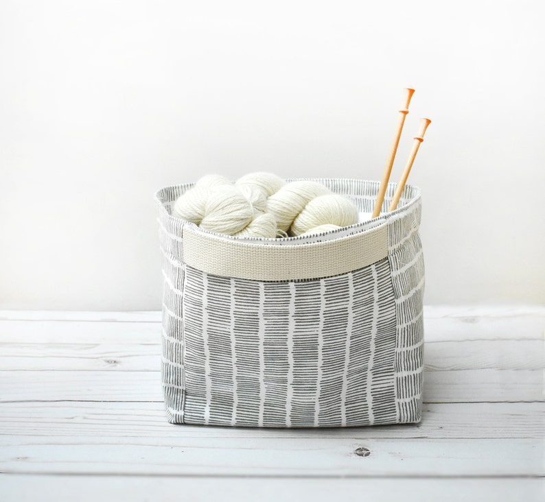 May include: A white fabric storage bin with a black and white geometric pattern. The bin is filled with white yarn balls and two wooden knitting needles.