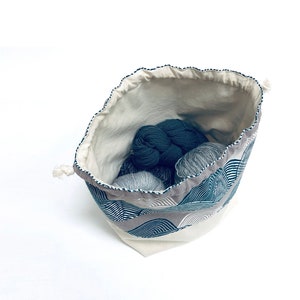 May include: A white drawstring bag with a blue and gray geometric pattern. The bag is filled with balls of yarn in shades of gray and blue.