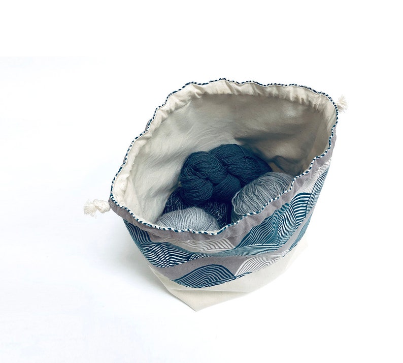 May include: A white drawstring bag with a blue and gray geometric pattern. The bag is filled with balls of yarn in shades of gray and blue.