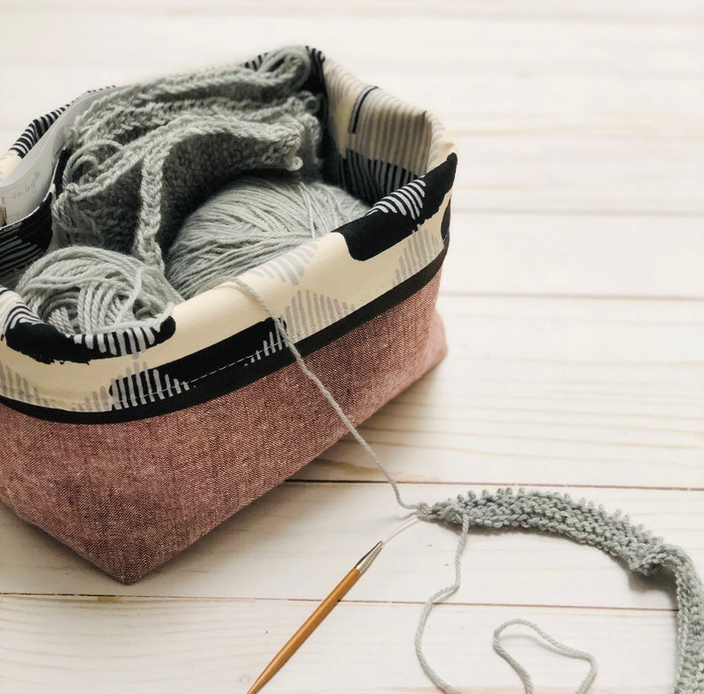 May include: A rectangular fabric basket with a pinkish-brown exterior and a black and white patterned interior. The basket contains gray yarn and knitted fabric. A wooden knitting needle is in the foreground, with a partially knitted piece of gray yarn.