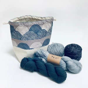 May include: A gray and white fabric storage bag with a drawstring closure, filled with blue and gray yarn skeins. The bag has a pattern of overlapping arches.