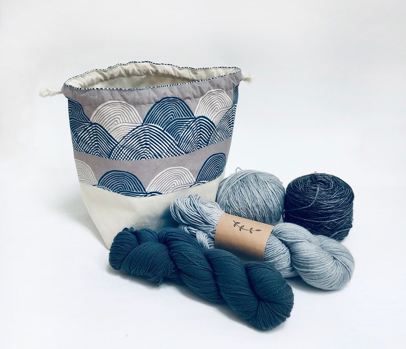 May include: A gray and white fabric storage bag with a drawstring closure, filled with blue and gray yarn skeins. The bag has a pattern of overlapping arches.