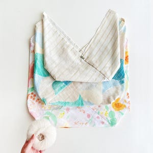 May include: A folded tote bag with a striped top and floral patterned body. The bag is layered over a floral patterned fabric. A ball of white yarn is held in a hand near the bag.