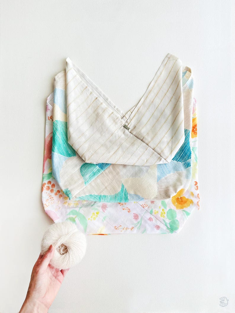 May include: A folded tote bag with a striped top and floral patterned body. The bag is layered over a floral patterned fabric. A ball of white yarn is held in a hand near the bag.
