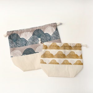May include: Two drawstring bags with a white background and a pattern of stylized mountains. One bag has a blue and gray mountain pattern, and the other has a gold and white mountain pattern.