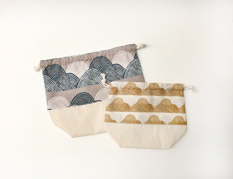 May include: Two drawstring bags with a white background and a pattern of stylized mountains. One bag has a blue and gray mountain pattern, and the other has a gold and white mountain pattern.