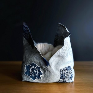 May include: A handmade fabric basket with a patchwork design. The basket is gray with navy blue accents and floral and geometric patterns. The interior is filled with white yarn. The basket is sitting on a wooden surface.