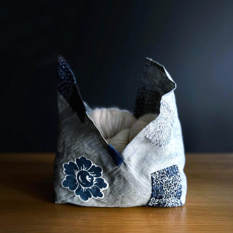 May include: A handmade fabric basket with a patchwork design. The basket is gray with navy blue accents and floral and geometric patterns. The interior is filled with white yarn. The basket is sitting on a wooden surface.
