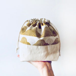 May include: A white drawstring bag with a gold and white patterned fabric lining. The bag is made of a soft, natural fabric and has a simple, elegant design.