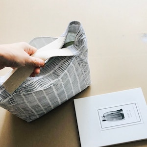 May include: A white tote bag with a black and white geometric pattern. The bag has a cream-coloured strap and is partially open, revealing the inside of the bag. The bag is sitting on a light brown surface. A white booklet with the text "Minette" is visible in the background.