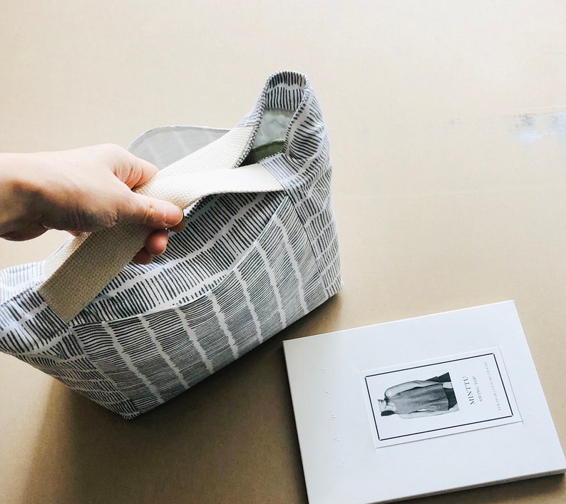 May include: A white tote bag with a black and white geometric pattern. The bag has a cream-coloured strap and is partially open, revealing the inside of the bag. The bag is sitting on a light brown surface. A white booklet with the text "Minette" is visible in the background.
