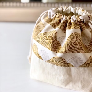 May include: A white drawstring bag with a gold and white geometric pattern. The bag is made of cotton fabric and has a simple design.
