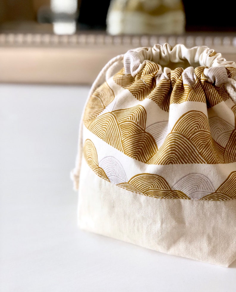 May include: A white drawstring bag with a gold and white geometric pattern. The bag is made of cotton fabric and has a simple design.