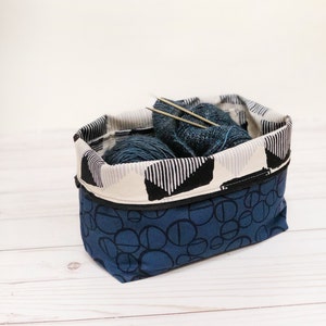 May include: A blue fabric basket with a black and white patterned trim, containing blue yarn and knitting needles. The basket's exterior features a dark blue pattern of overlapping circles. The basket is designed for storing craft supplies.