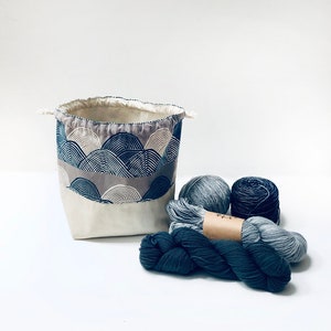 Puede incluir: Una bolsa de almacenamiento de tela blanca con cierre de cord&oacute;n y un patr&oacute;n geom&eacute;trico de ondas azules y grises. La bolsa est&aacute; llena de ovillos de lana azul.