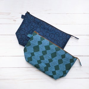 May include: Two rectangular zippered pouches. One is navy blue with a black circle pattern. The other is teal with a green diamond pattern. Both have brown zippers and are on a white wooden surface.