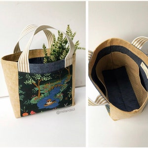 May include: A beige tote bag with a white and brown striped handle. The bag is lined with a dark blue fabric with white polka dots. The exterior of the bag features a colourful fabric with a nature scene.