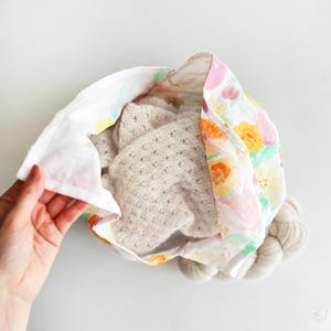 May include: A floral fabric bag with a light-colored knitted item inside. The bag's fabric features a pattern of colorful flowers. A skein of yarn is visible at the bottom of the bag. The bag is being held open, revealing its contents.