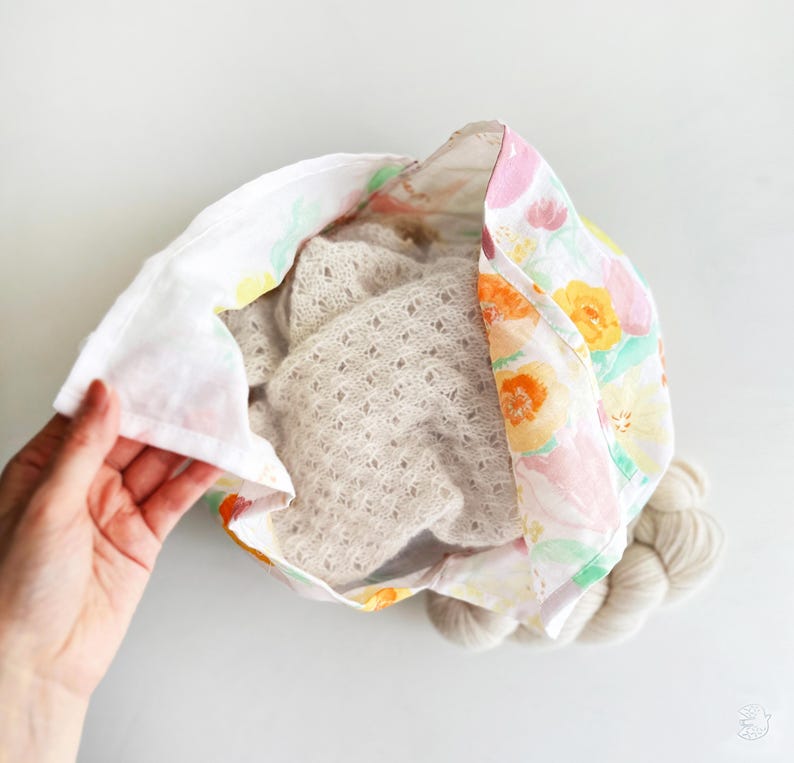 May include: A floral fabric bag with a light-colored knitted item inside. The bag's fabric features a pattern of colorful flowers. A skein of yarn is visible at the bottom of the bag. The bag is being held open, revealing its contents.