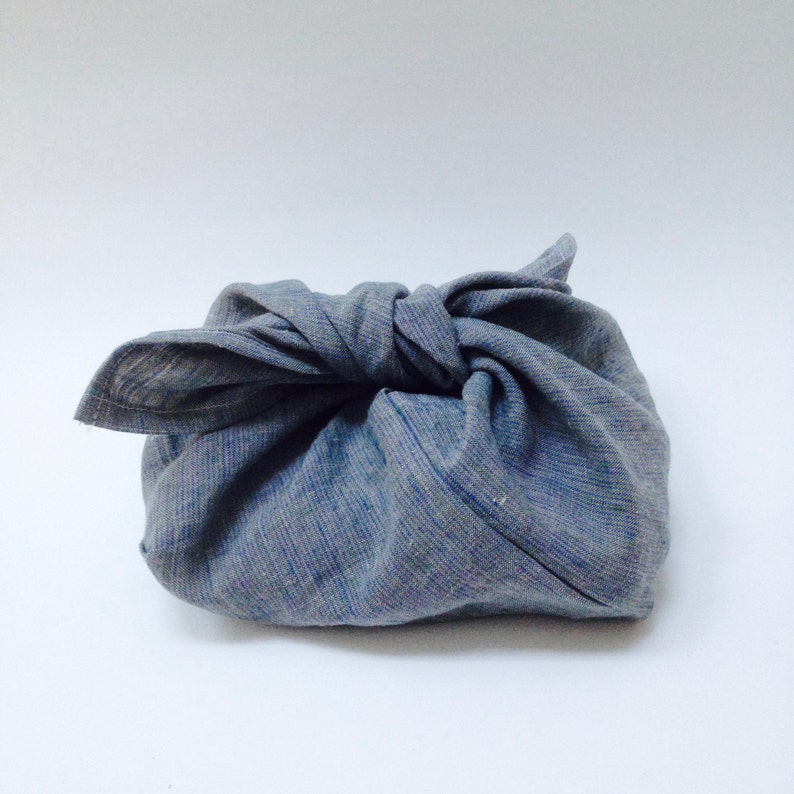 May include: A gray fabric furoshiki cloth, tied in a knot. The cloth is a muted gray color with subtle blue threads woven throughout. The knot is centered, creating a compact, wrapped appearance. The background is plain white.