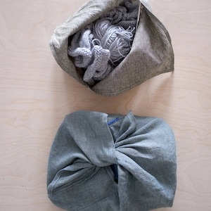 May include: Two gray linen bags on a light wood surface. One bag is open, filled with gray yarn and knitted items. The other bag is closed, tied in a knot. The bags are likely for storing or carrying craft supplies.