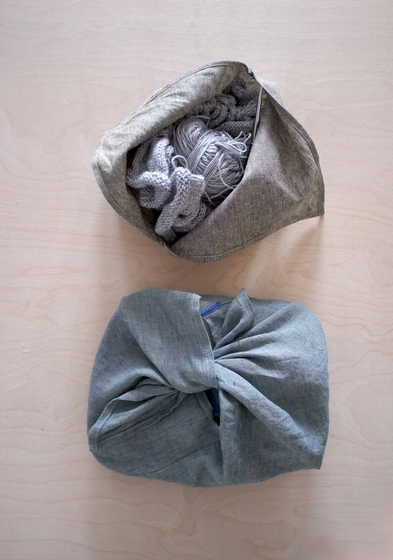 May include: Two gray linen bags on a light wood surface. One bag is open, filled with gray yarn and knitted items. The other bag is closed, tied in a knot. The bags are likely for storing or carrying craft supplies.