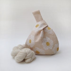 Puede incluir: Una bolsa de tela beige con un patr&oacute;n de margaritas blancas y centros amarillos. La bolsa est&aacute; anudada en la parte superior y tiene un solo asa. Un ovillo de hilo blanco est&aacute; en primer plano.