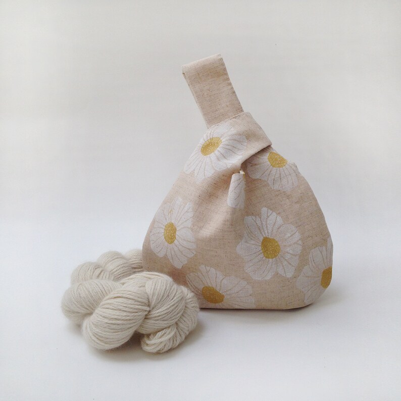 Puede incluir: Una bolsa de tela beige con un patr&oacute;n de margaritas blancas y centros amarillos. La bolsa est&aacute; anudada en la parte superior y tiene un solo asa. Un ovillo de hilo blanco est&aacute; en primer plano.